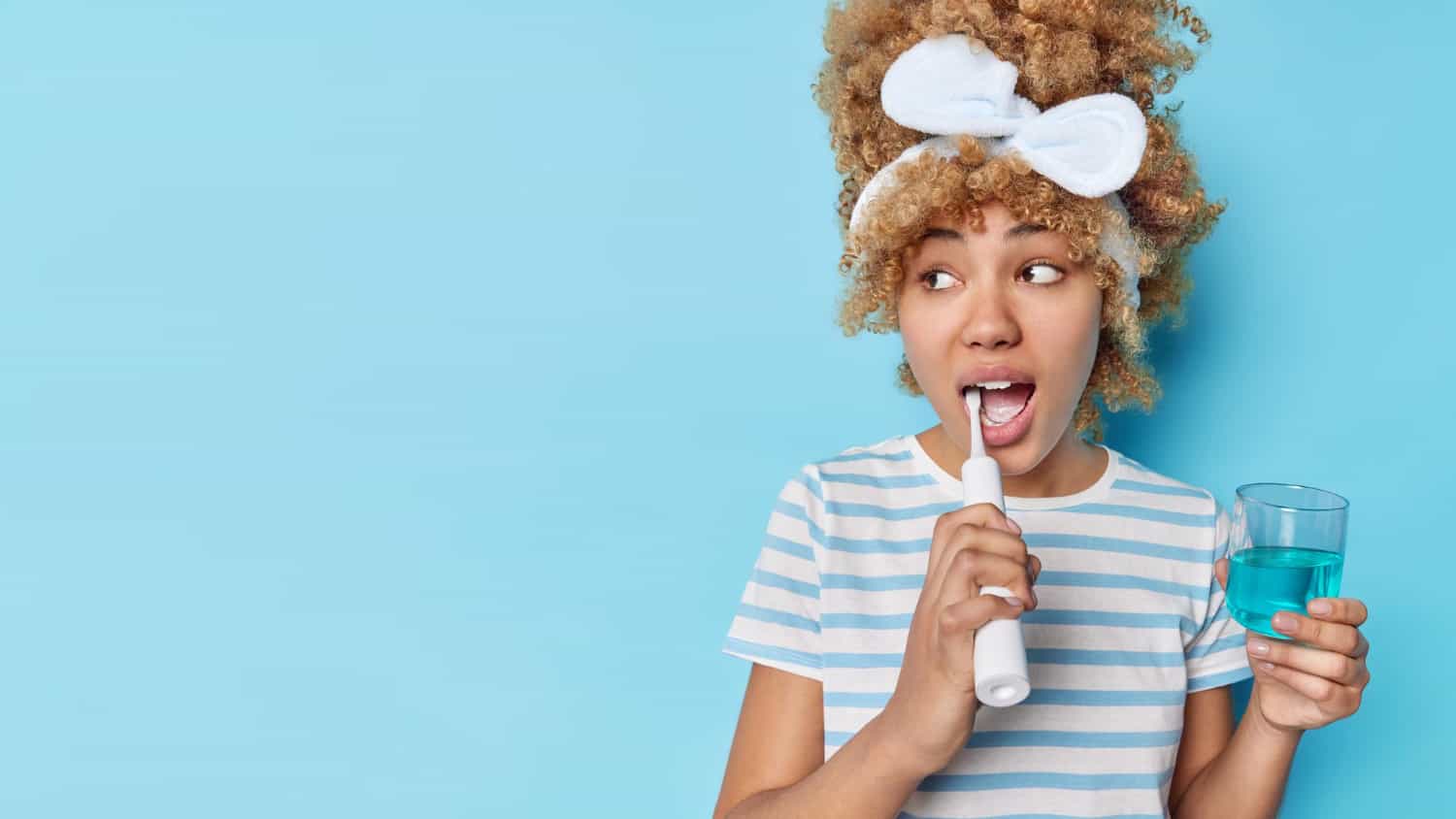 A young woman with curly hair is shown brushing her teeth with an electric toothbrush while holding a glass of blue mouthwash, demonstrating a healthy daily oral hygiene routine encouraged by ZPD.
